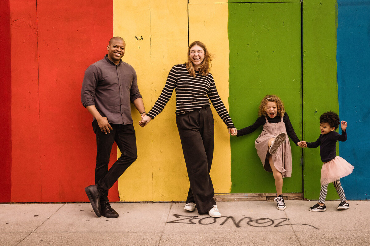Mixed race San Francisco family holds hand and stands in front of color block rainbow wall