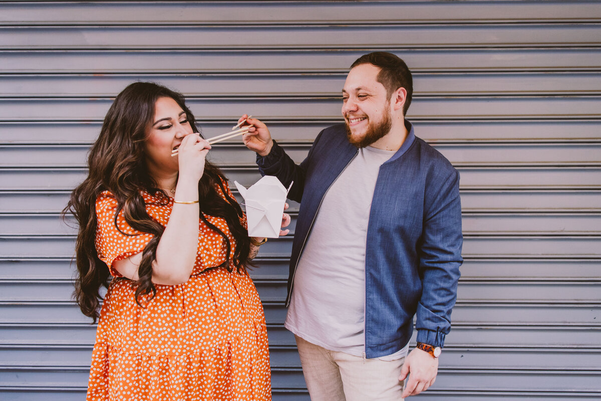 A couple laughing and eating boxed chinese food in front of a garage door at chinatown in nyc during their engagement shoot