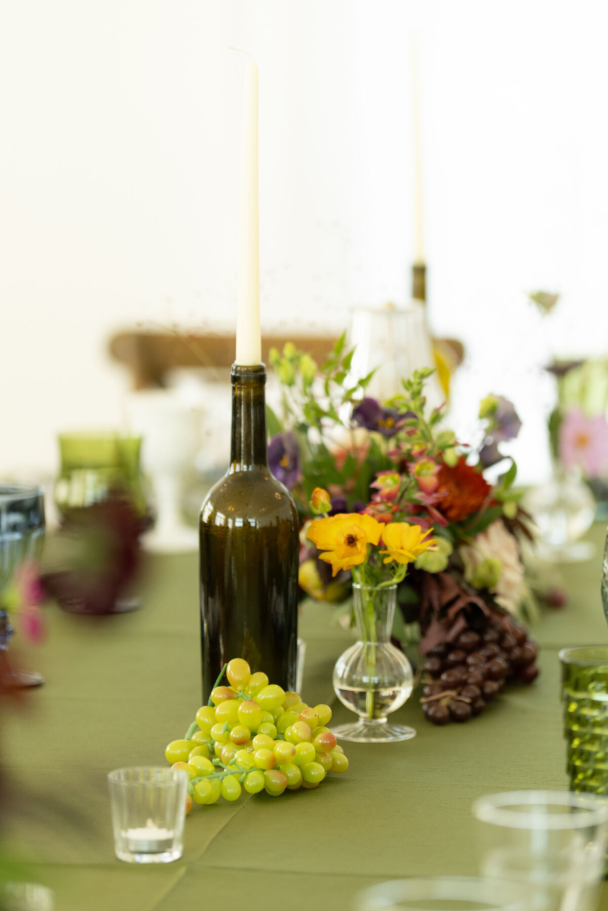 Wedding reception table decor featuring a wine bottle taper candle, green grapes, and bright garden-style floral arrangements in a modern, organic design.
