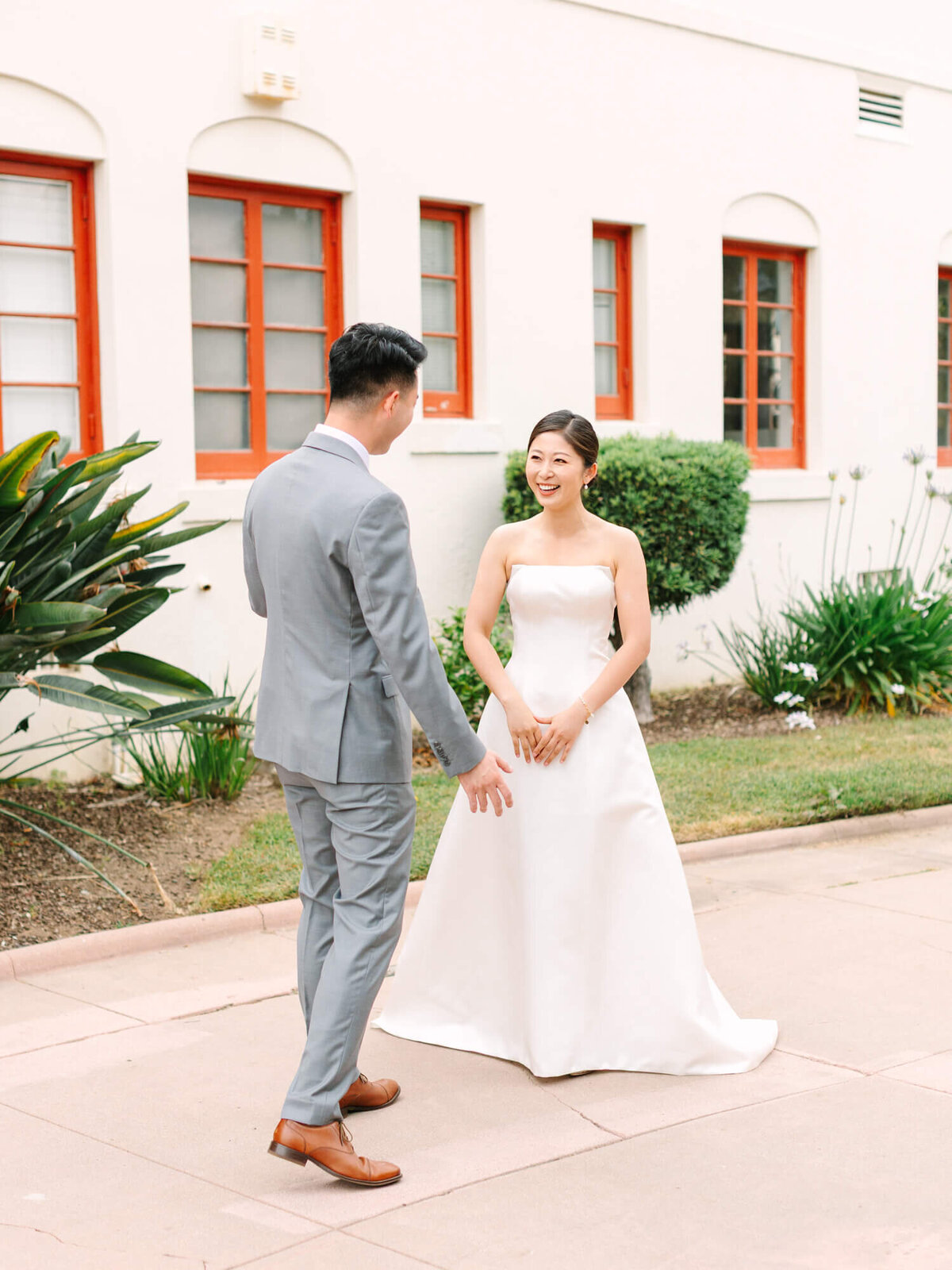 A bride in a white gown and a groom in a gray suit joyfully face each other outdoors. Red-framed windows and greenery add charm to the scene.