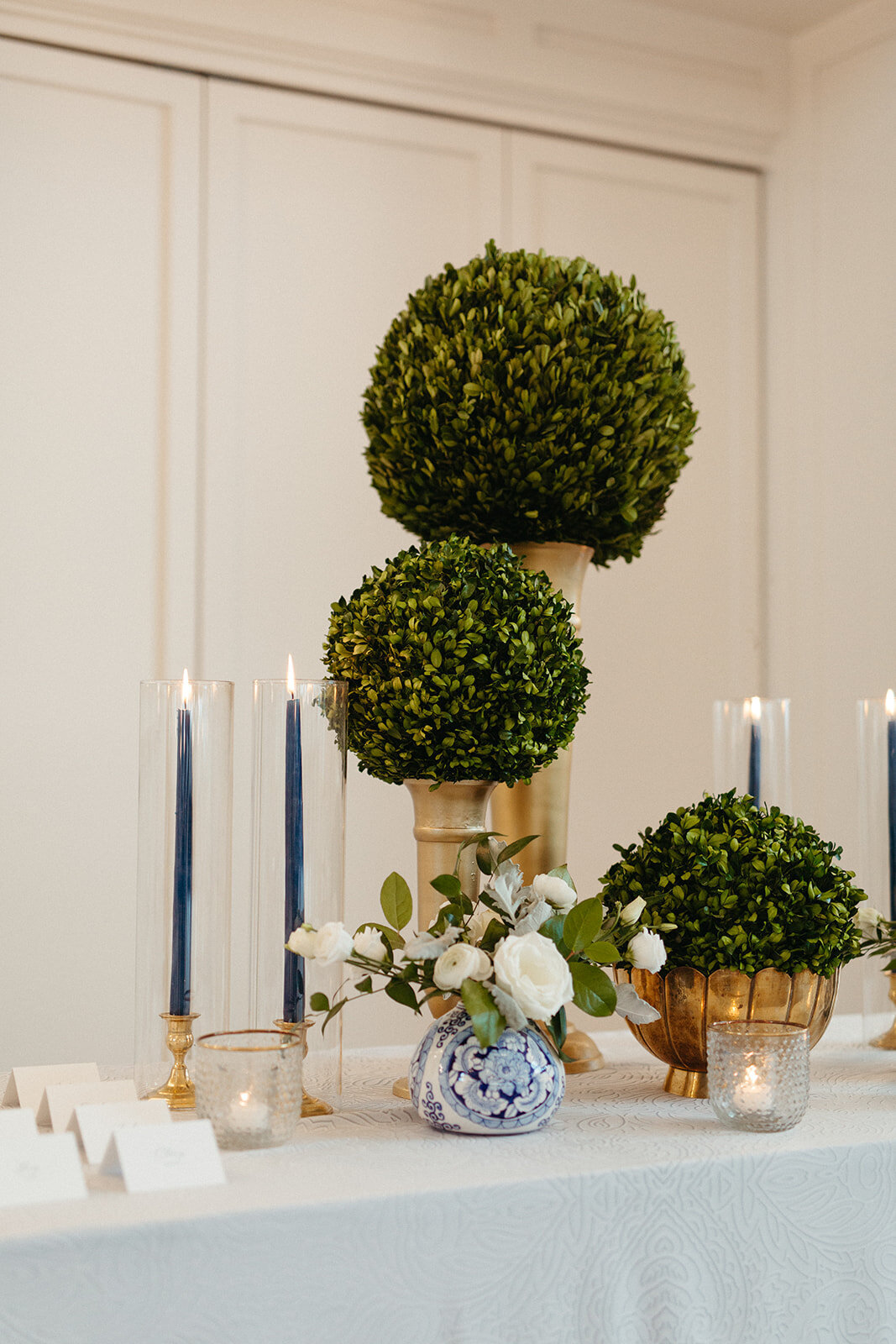 White place cards with flower arrangement, lit candles and plants in gold stands atop a table with white linen