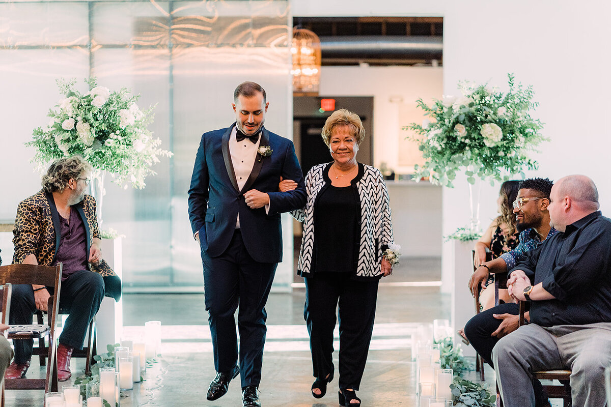 Groom walks down the aisle arm-in-arm with a smiling woman, possibly a family member, as guests watch with warm smiles.