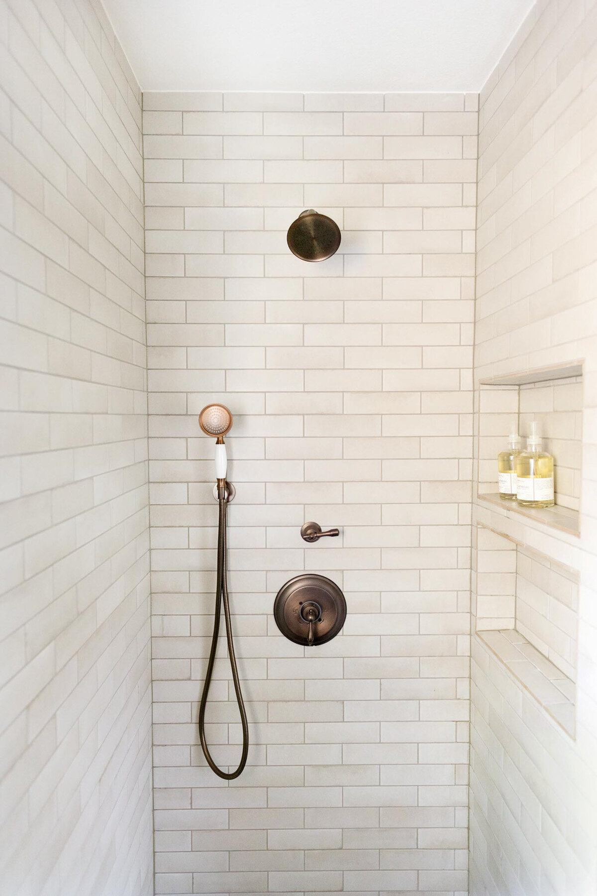 View of shower facing the shower head with off white subway tiles. Antique bronze shower fixtures with two valves, a shower head, and a handheld shower. Two niches on the wall to the right with soap bottles.