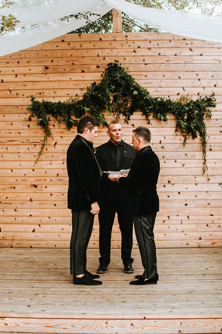 Two grooms wearing black tuxedos exchange vows in front of an officiator on a wooden stage outdoors.