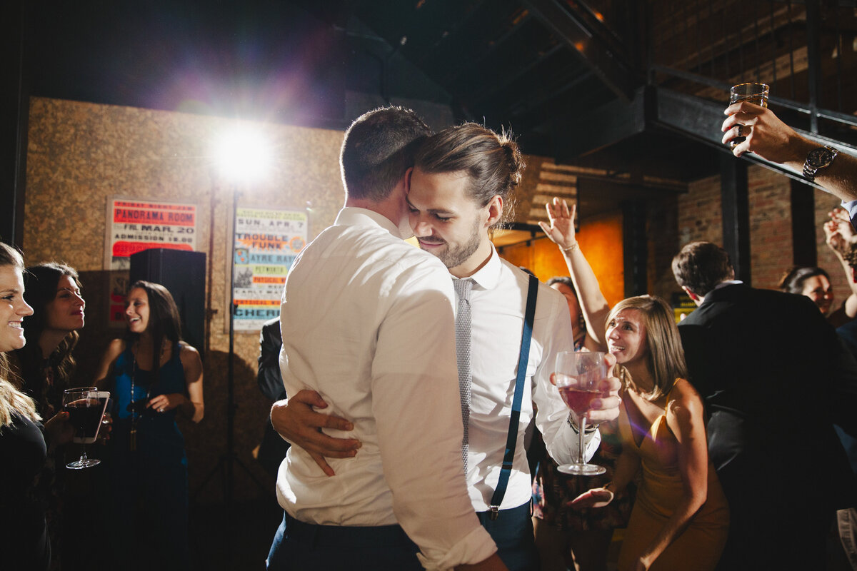 A newlywed couple standing close together and dancing with guests cheering behind them 