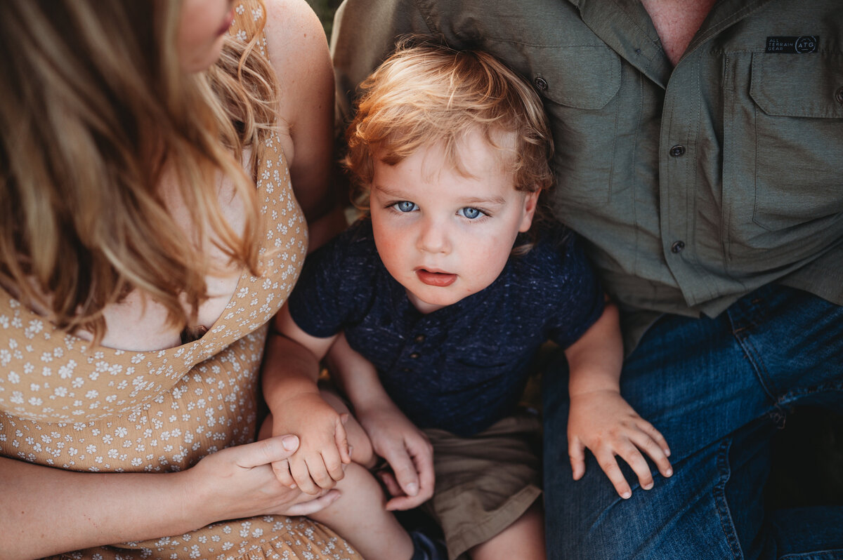 Little blonde boy with blue eyes gazes at the camera while sitting with his mother and father