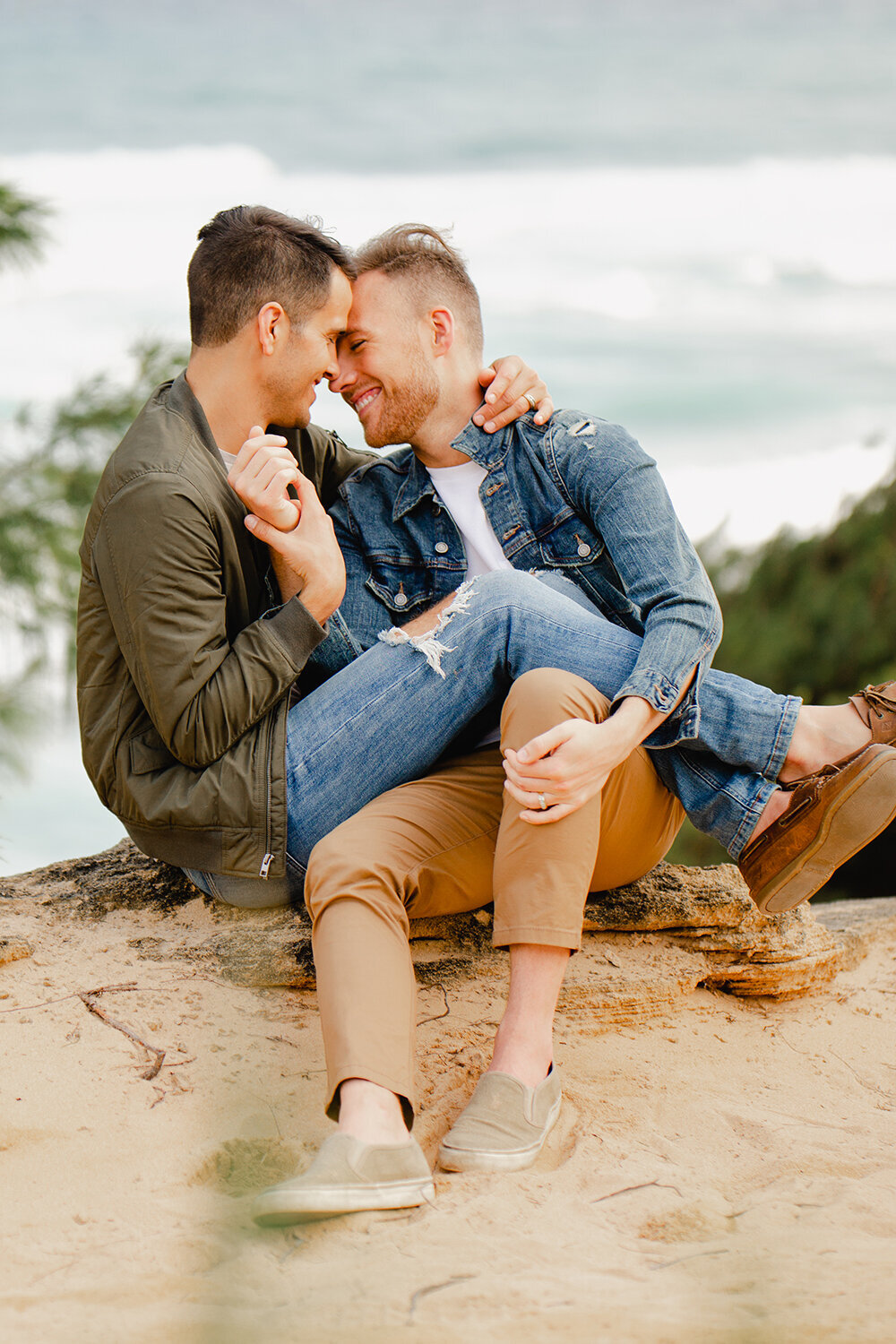 LGBT couples session on a secluded beach in Hawaii. A man sits on the beach with his boyfriend's legs wrapped around him.