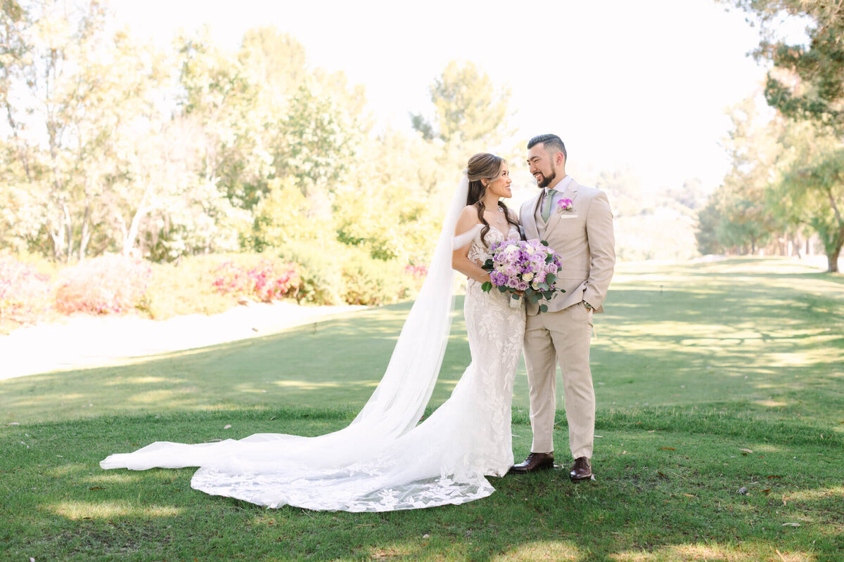 A bride and groom stand smiling on a sunny lawn. The bride wears a white lace gown with a long veil, holding a bouquet of purple flowers. The groom is in a tan suit with a purple boutonniere. Trees and greenery surround them, creating a serene, romantic setting at Coyote Hills Golf Course.