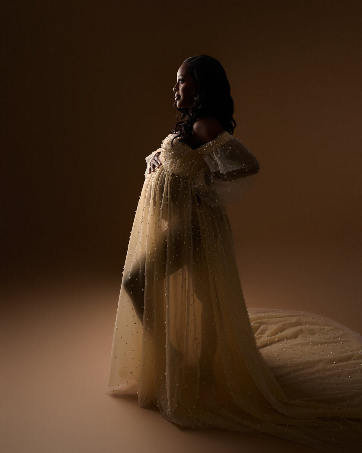Backlit studio maternity image in sheer golden gown with soft shadows in Austin, TX