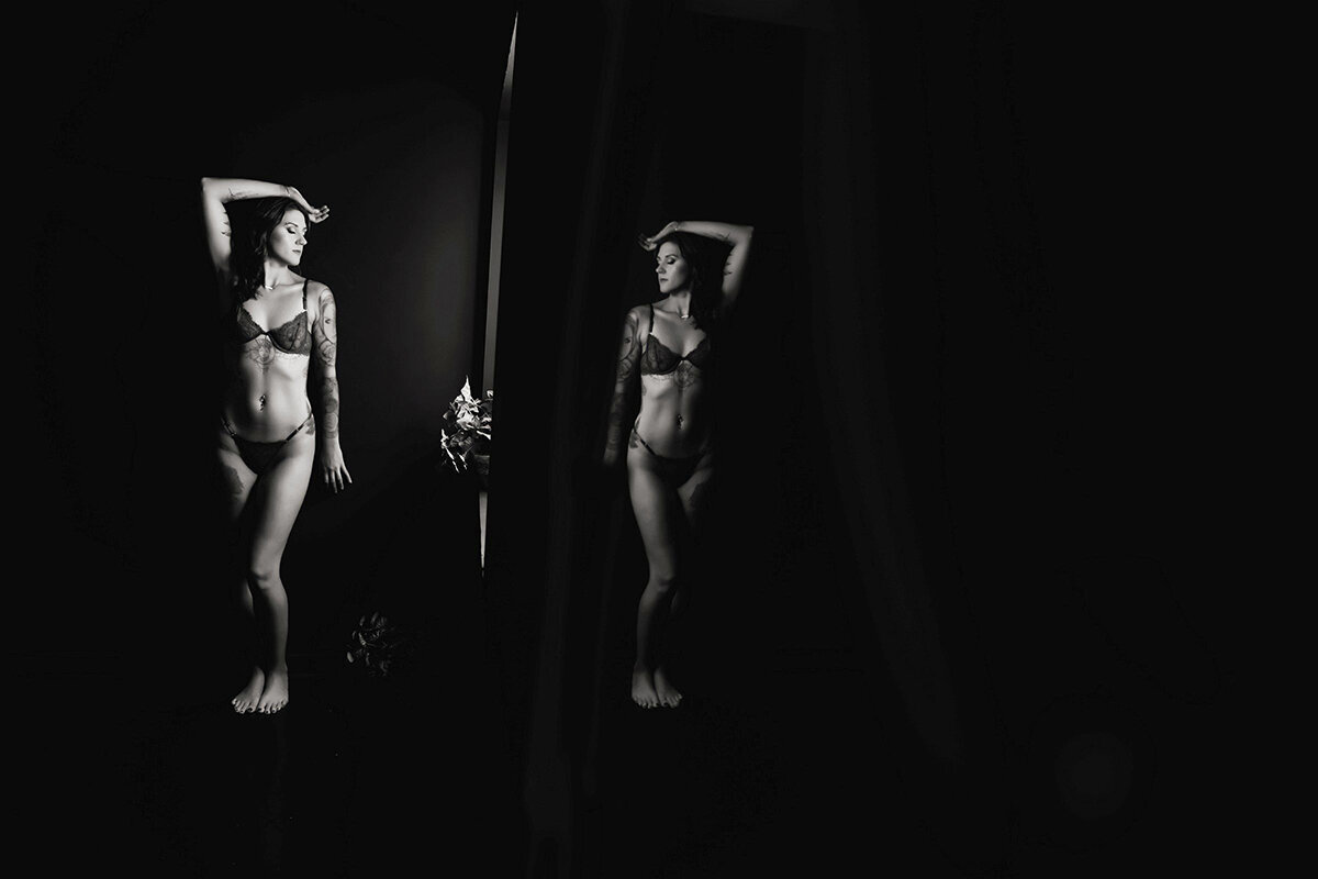 A woman in lingerie poses confidently against a dark backdrop, her reflection visible in a mirror. The mood is elegant, with soft lighting accentuating her form.