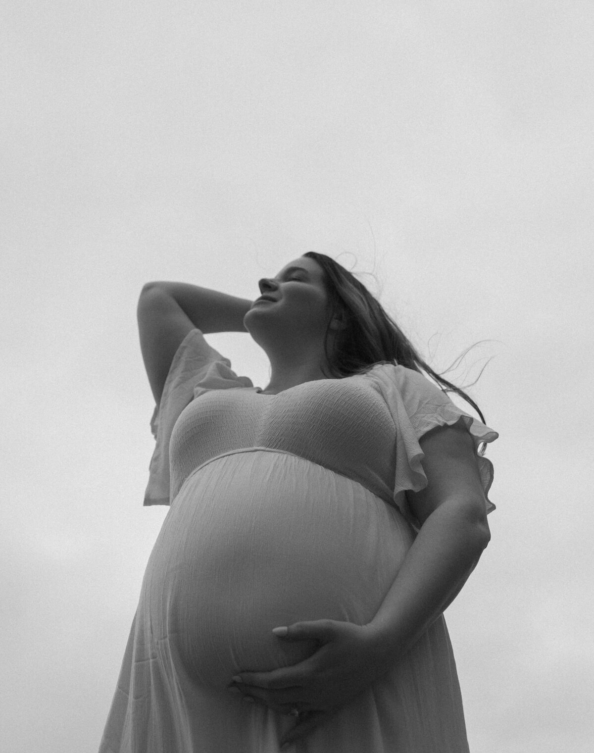 pregnant woman wearing a white dress holds her belly and stands against a cloudy sky backdrop at Oregon Coast