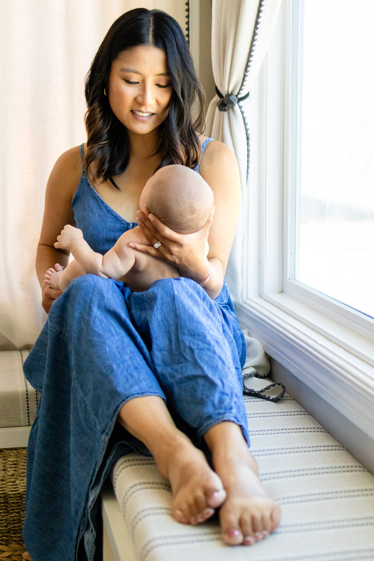 Mother smiling at baby while sitting by a bright window, photographed by San Francisco newborn photographer.