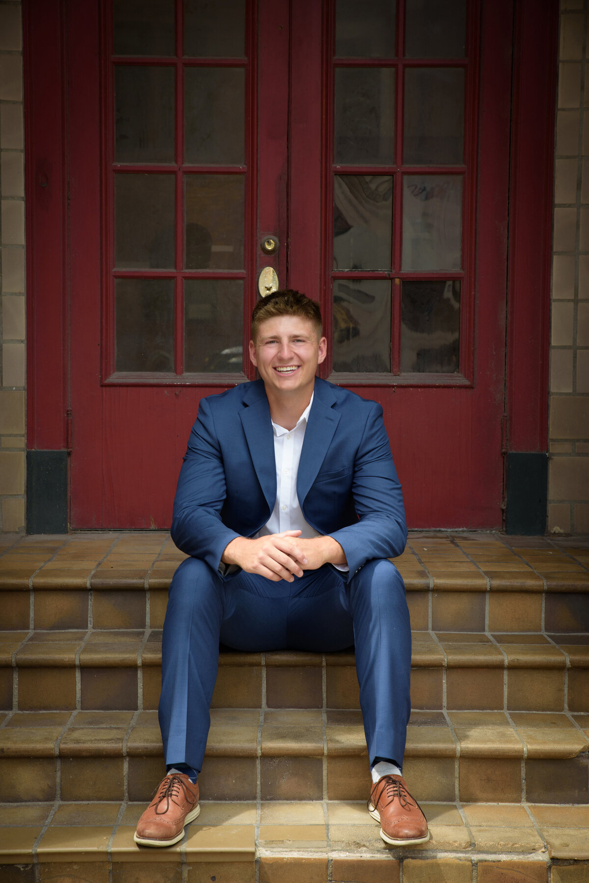De Pere High School senior boy wearing a blue suit with white shirt sitting on stairs in front of red door in urban setting in Downtown Green Bay, Wisconsin