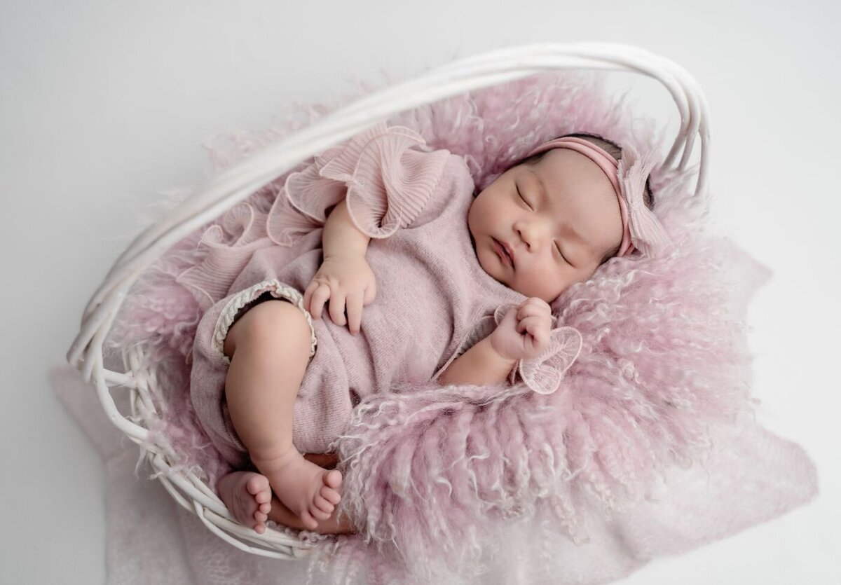 Baby in pink basket close-up – Newborn baby girl swaddled in pink with a matching headband, sleeping soundly in a white wicker basket lined with a soft pink fur blanket.
