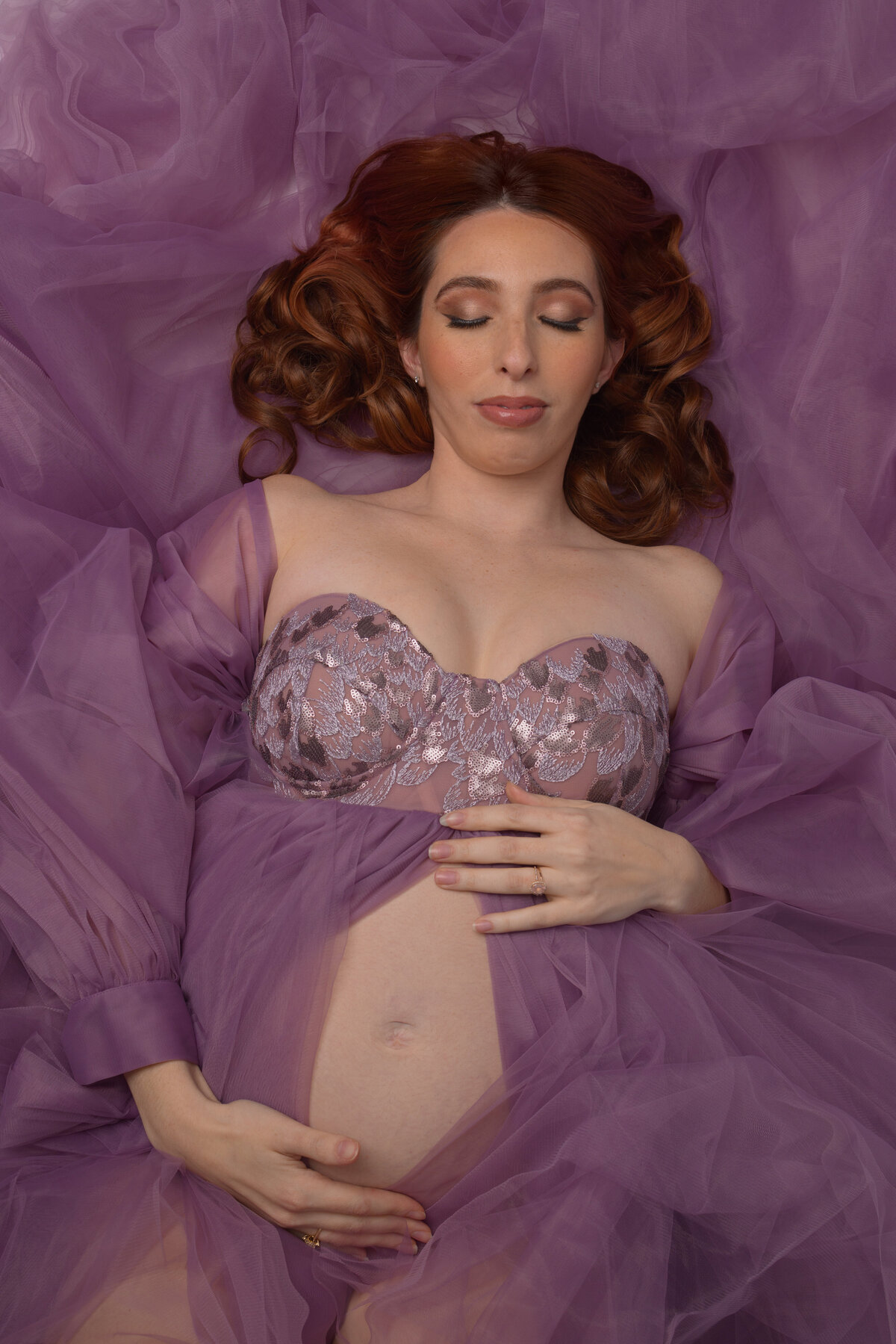 Elegant maternity portrait with flowing purple tulle fabric and sparkly bralette surrounded by dreamy violet lighting.