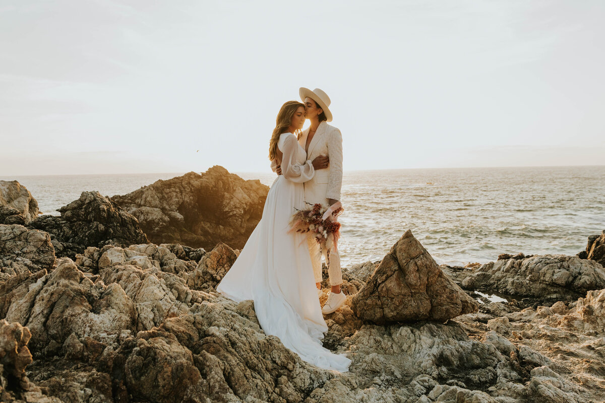 two brides standing on rocks next to ocean while embracing