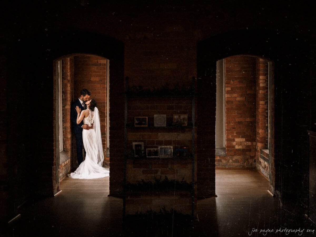 A wedding couple kissing in the corner of a red brick room.