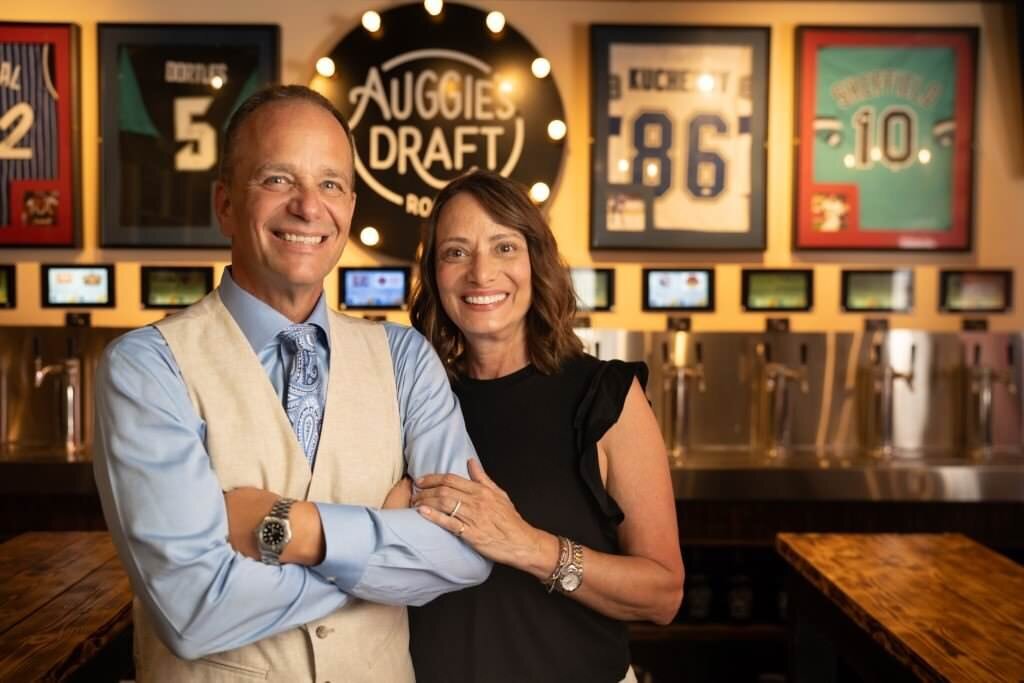 A-smiling-couple-poses-together-in-front-of-a-wall-decorated-with-sports-jerseys-at-Auggies-Draft-Room