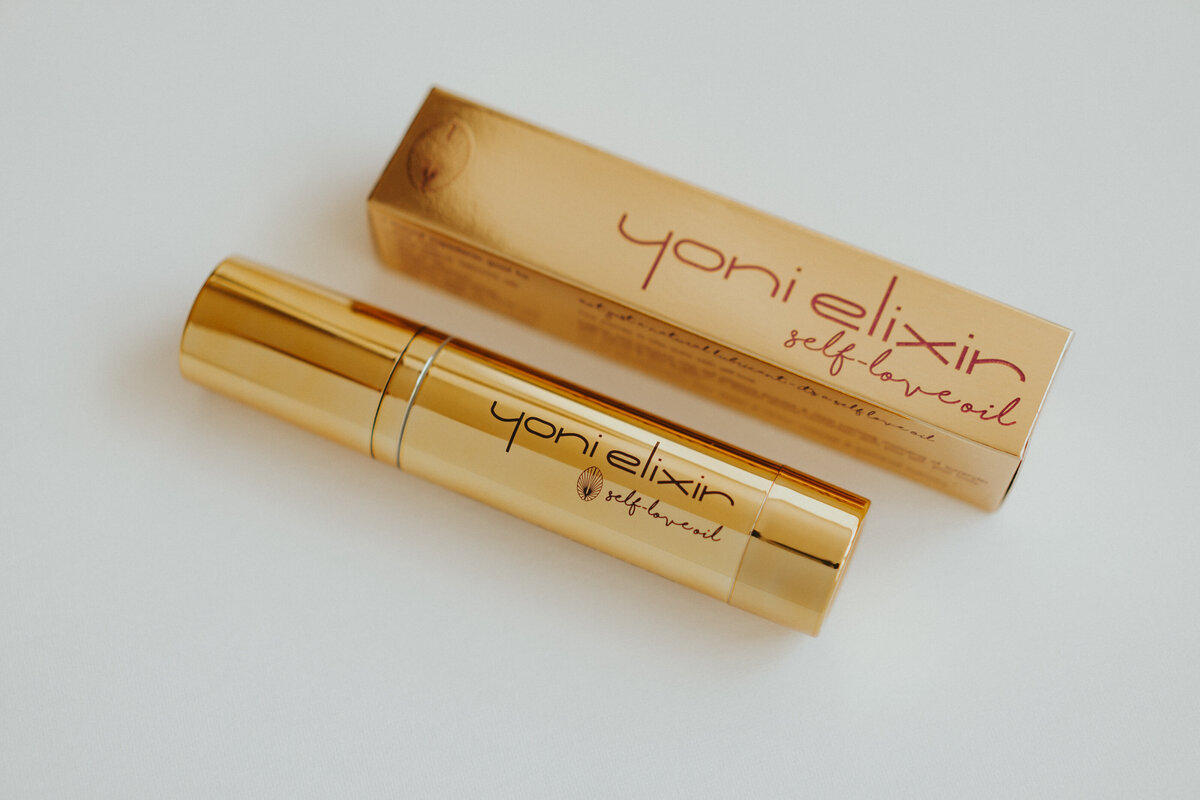 Yoni Elixir gold packaging displayed on desk, with sensual woman sitting behind