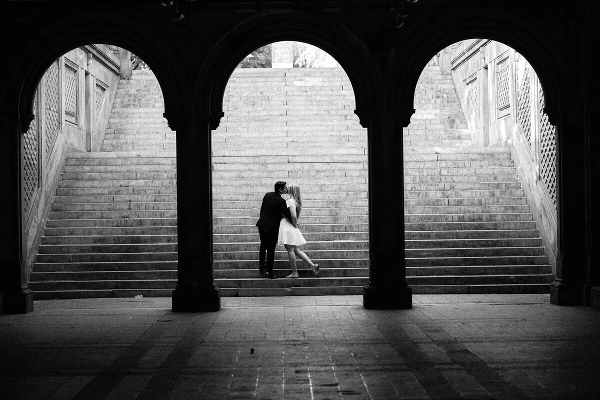 A couple embraces and kisses at the center of wide stone steps under an archway, framed by shadows, in a dramatic black and white setting captured by a talented film photographer NJ.