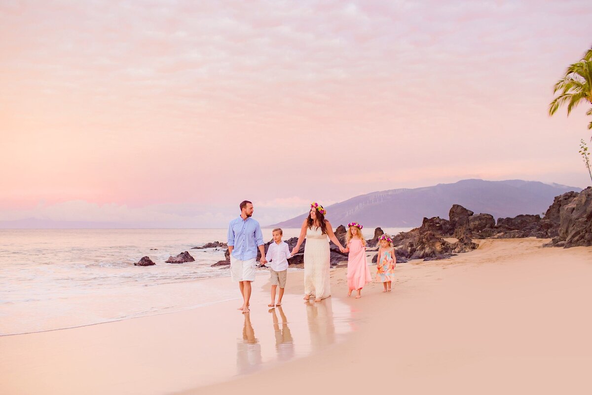 Family with three young kids walks hand-in-hand along the shoreline on Maui with mountains in the backdrop and beautiful pink sky by Love + Water