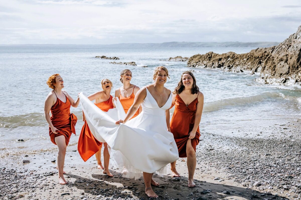 A bride and her bridesmaids on the beach walking away from the water with the bridesmaids holding the brides dress