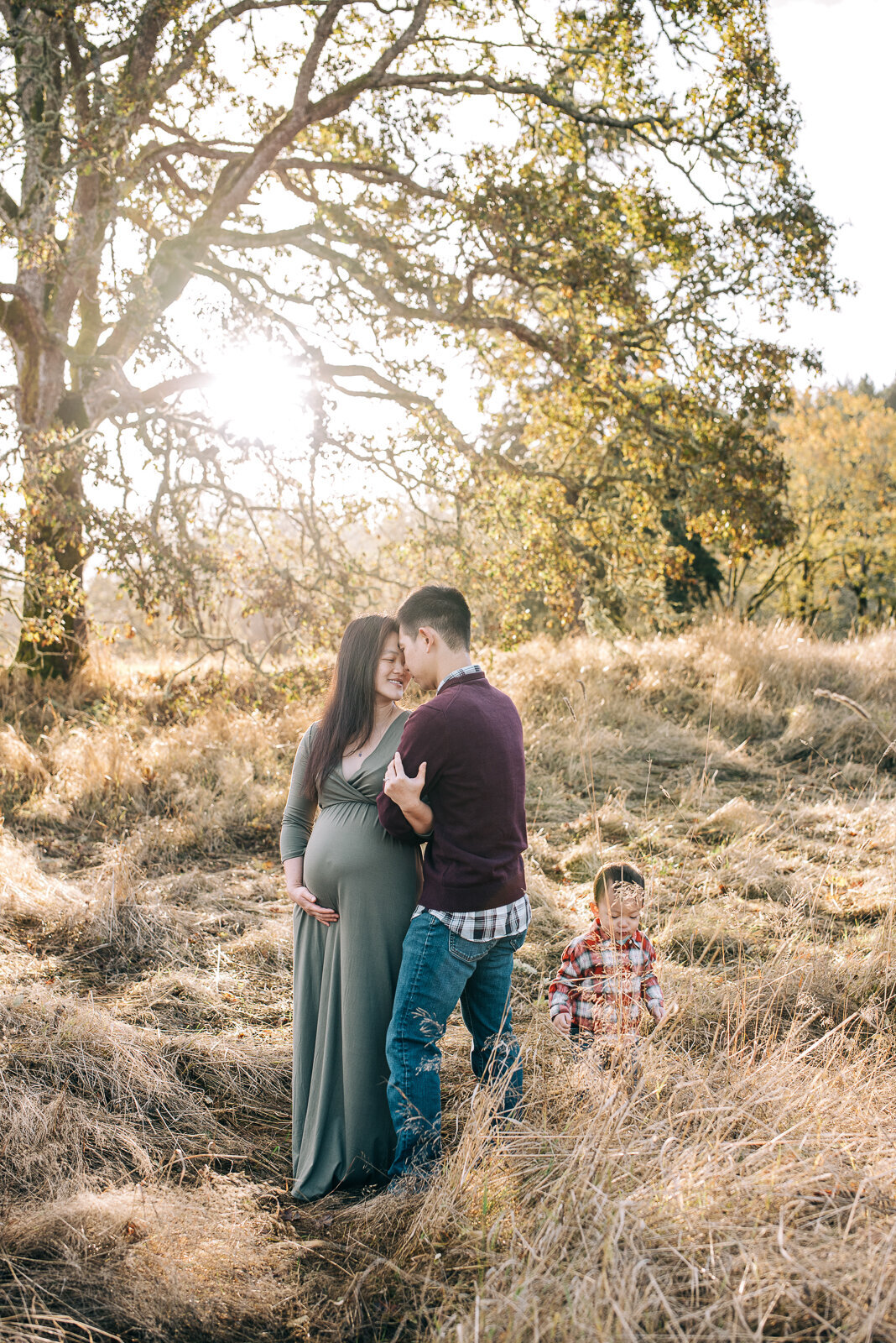 Husband standing behind his wife, wrapping his arms around her pregnant belly while their son plays at their feet nearby