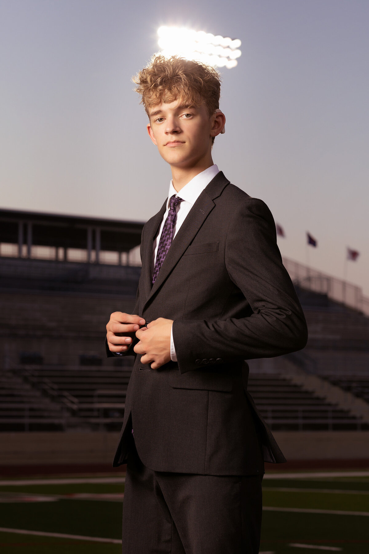 A senior in a dark suit stands confidently on the football field at Grapevine High School, adjusting his jacket with the stadium lights in the background. This Dallas senior photography captures his stylish look.