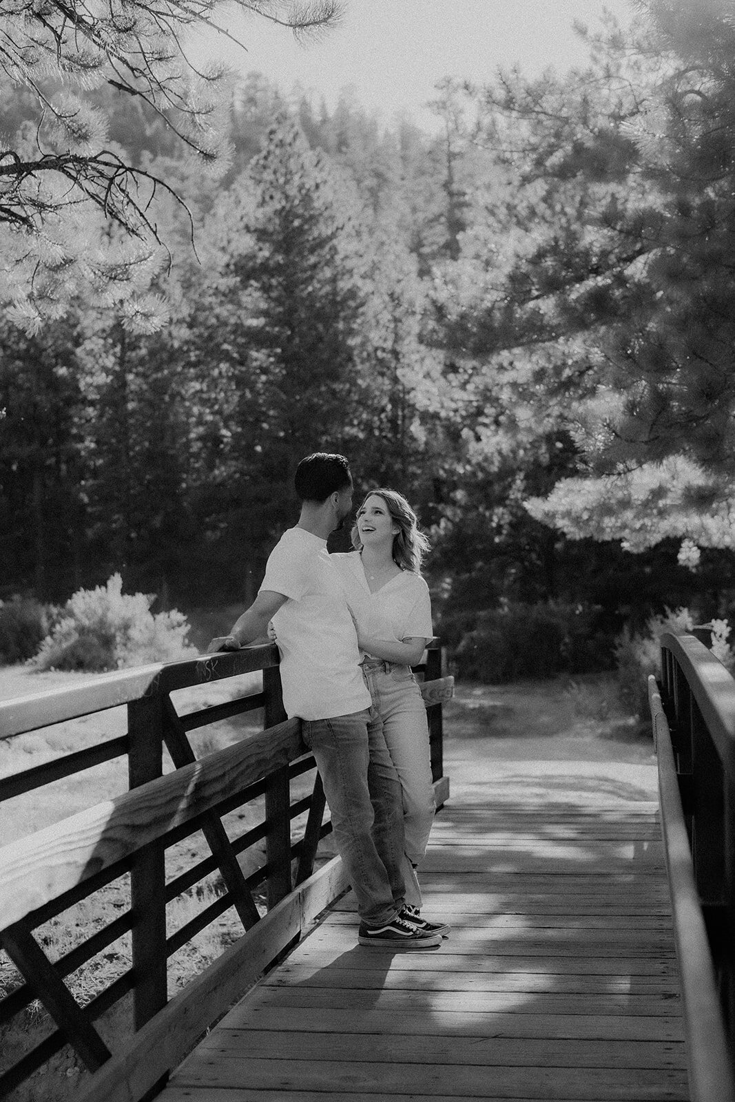 The couple stands on a bridge, laughing and sharing an intimate moment, with a forested backdrop bathed in soft light.
