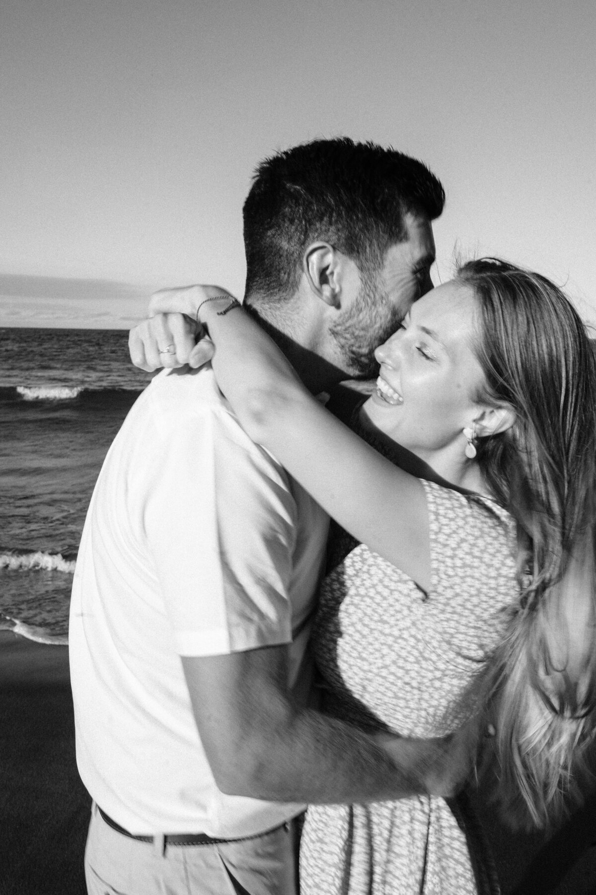 black and white photo of a couple hugging at laughing at taylors mistake beach in christchurch