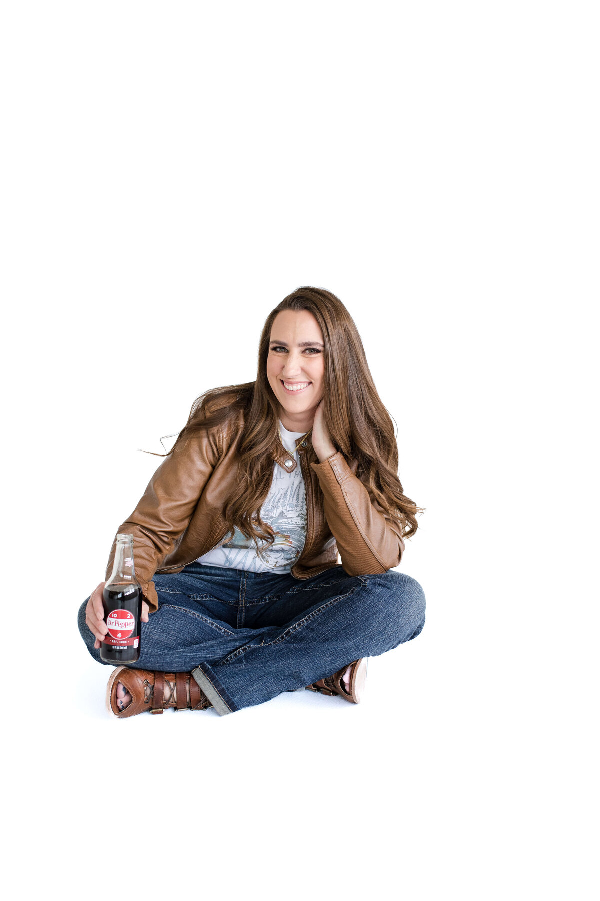brand photographer near me photographs brand photo with woman sitting criss cross while wearing jeans and a brown leather jacket holding a coke bottle and smiling as she rests her head on her hand
