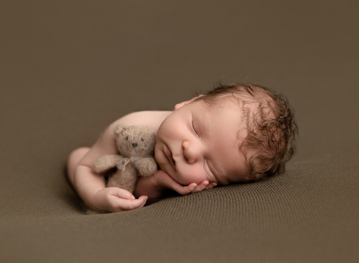 Newborn boy posed on a green backdrop facing the camer with his hand under his chin and clutching a tiny knitted bear