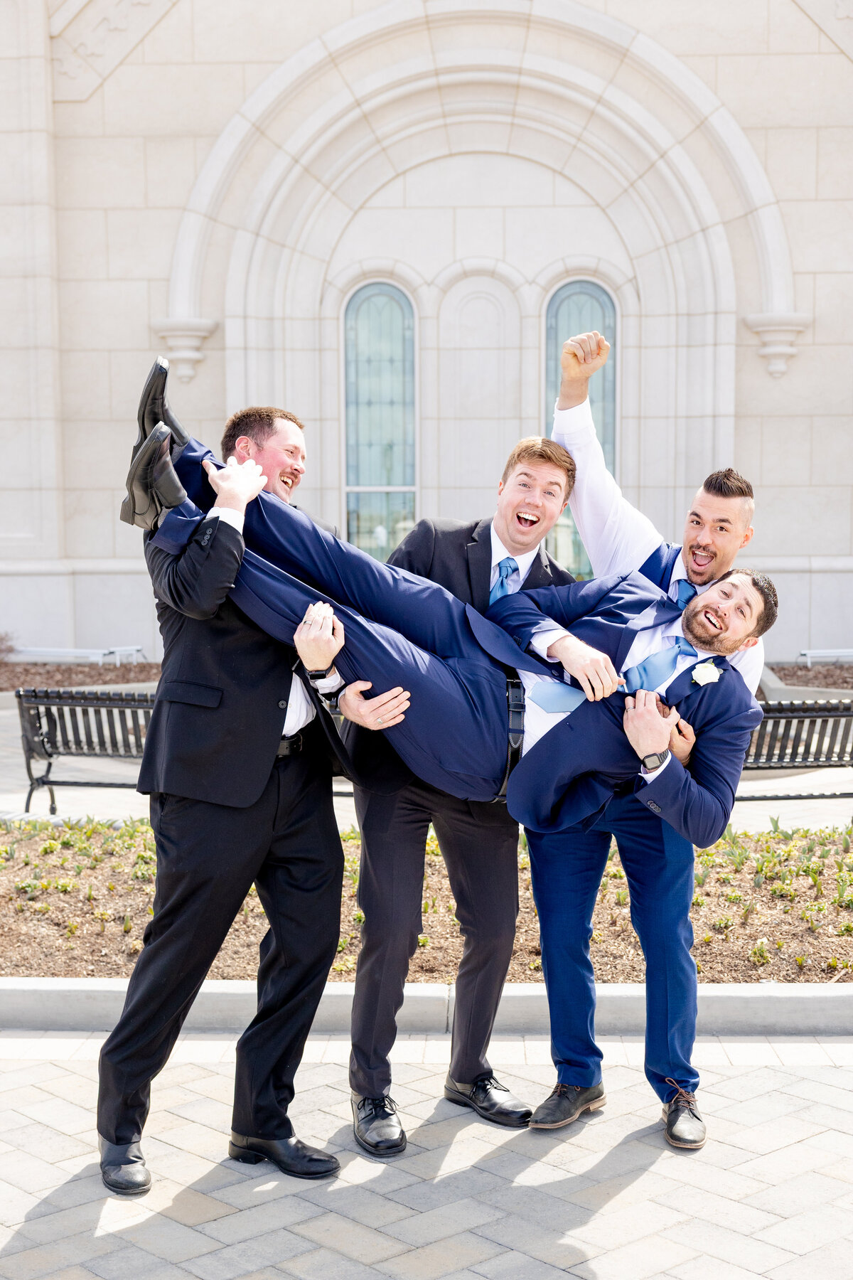 Four groomsmen cheerfully lift the groom in the air in front of the LDS temple, all smiling and laughing.