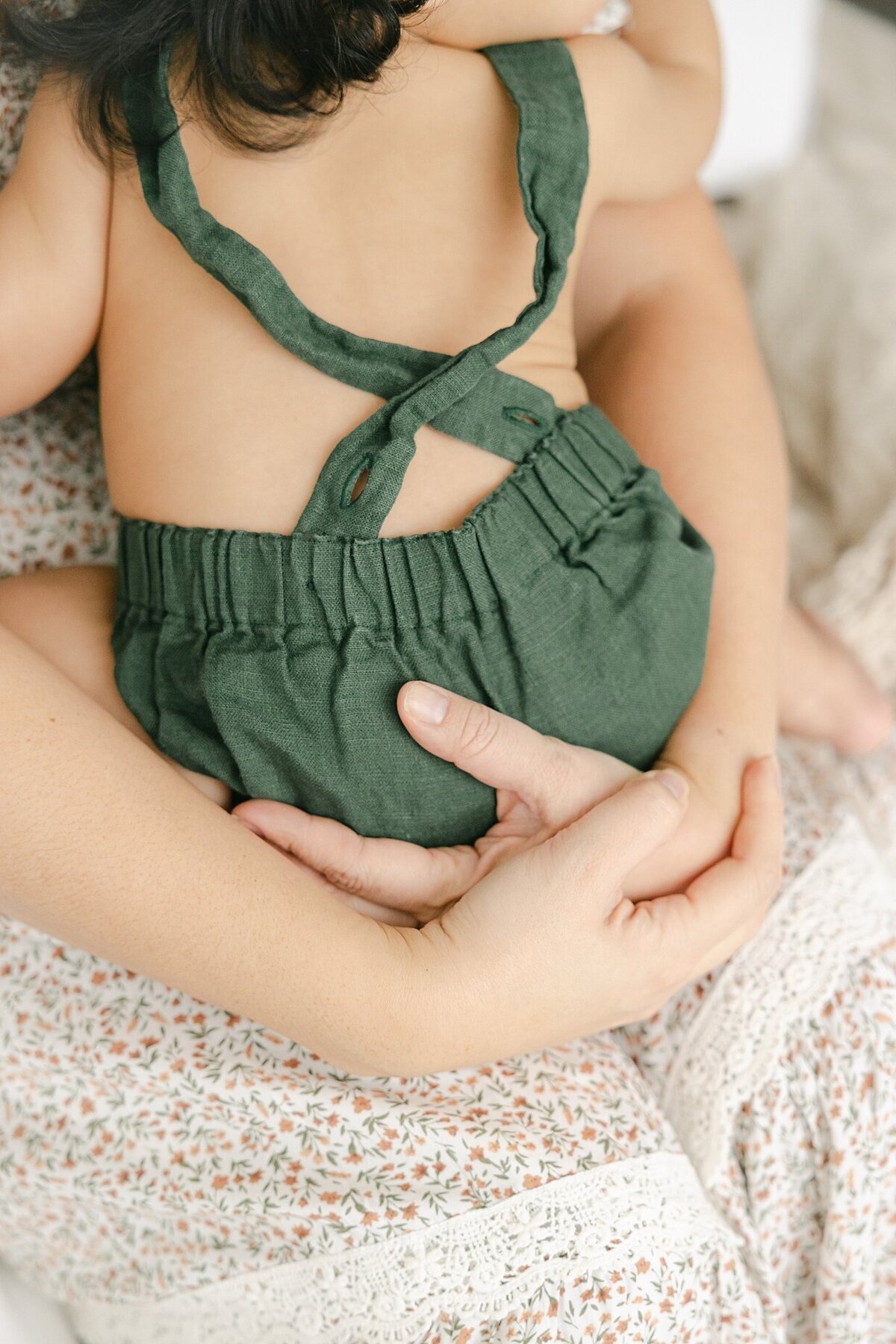 A close up of a mother in a floral dress holding her baby boy in green overalls on her lap as he hugs her during their Indianapolis family photography session