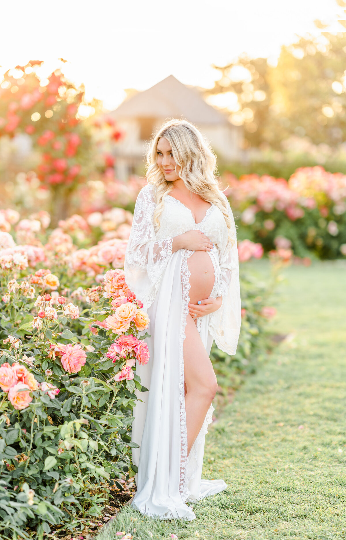 An expecting mother stands in a rose garden wearing a white gown holding and looking down at her beautiful baby bump photographed by Bay area photographer, Light Livin Photography.