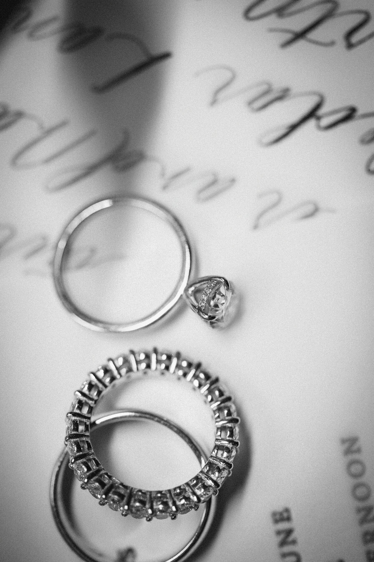 A close-up black and white image of three wedding rings arranged on top of a wedding invitation, with handwritten calligraphy softly blurred in the background and shadows creating contrast and texture across the paper.