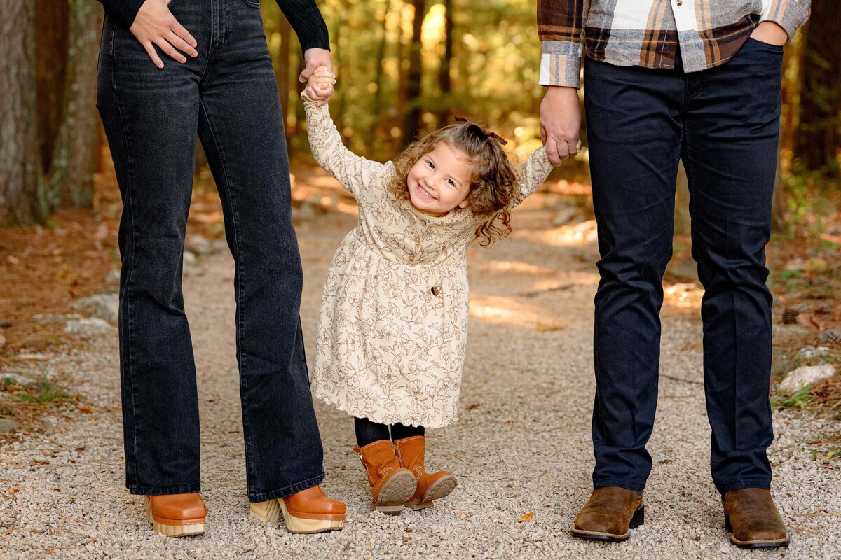 little girl with curly hair laughs and rocks back on her heels holding her parents hands