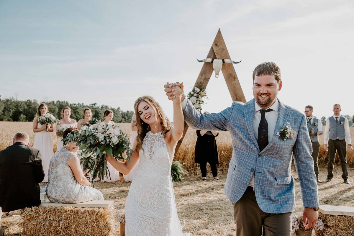 Bride and groom walking down the aisle at the end of their outdoor rustic farm ceremony. They are holding hands and raising their arms in the air. Guests are sitting on haybales.