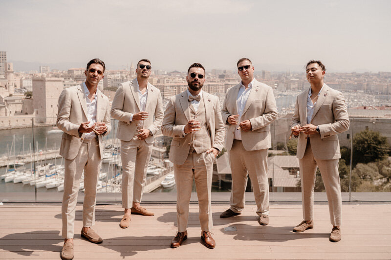 Le marié pose avec ses témoins en costume clair dans un style Vanity Fair, sur une terrasse avec vue sur le port de Marseille.