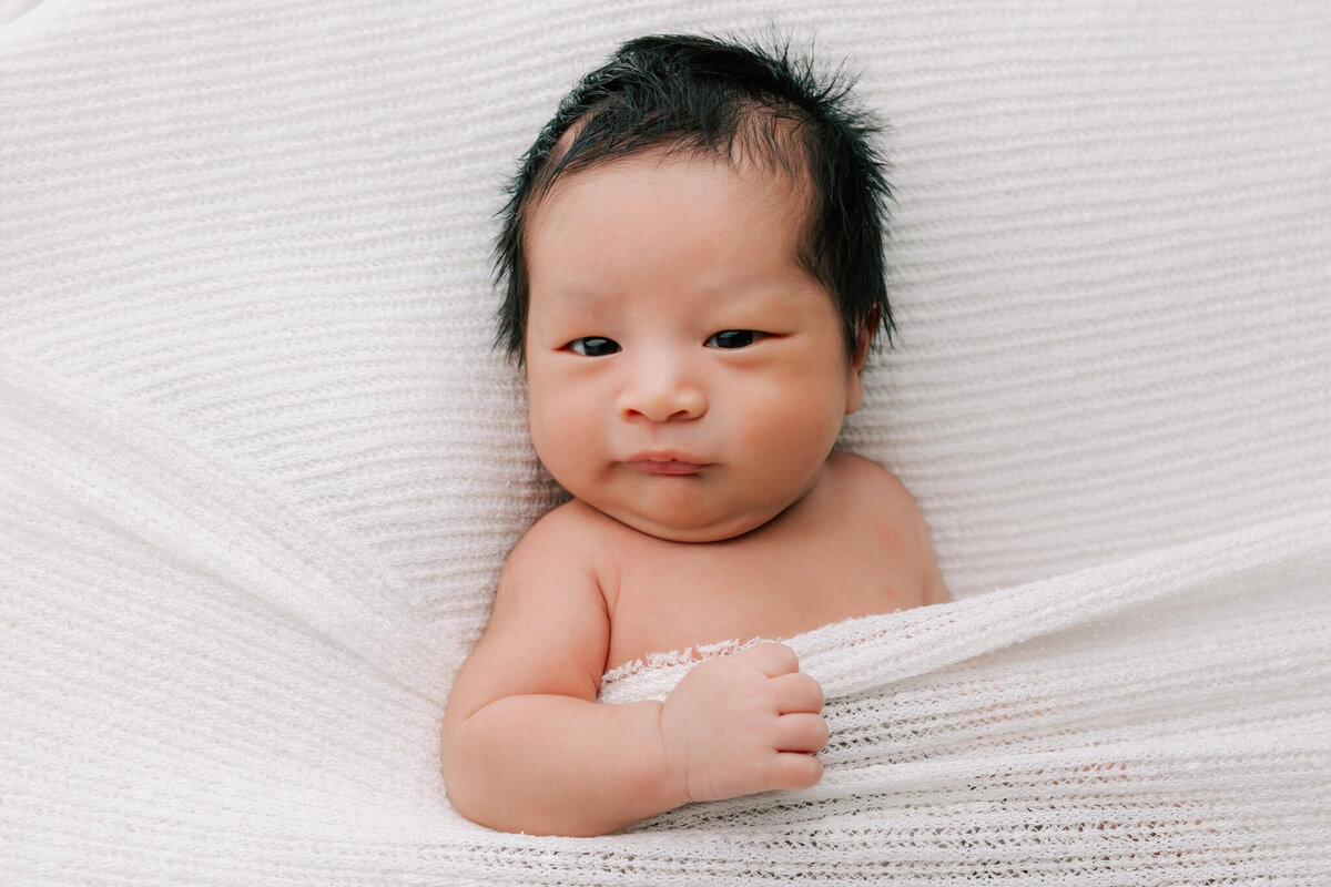 portrait of newborn boy with black hair laying on white blanket with his arm over the blanket