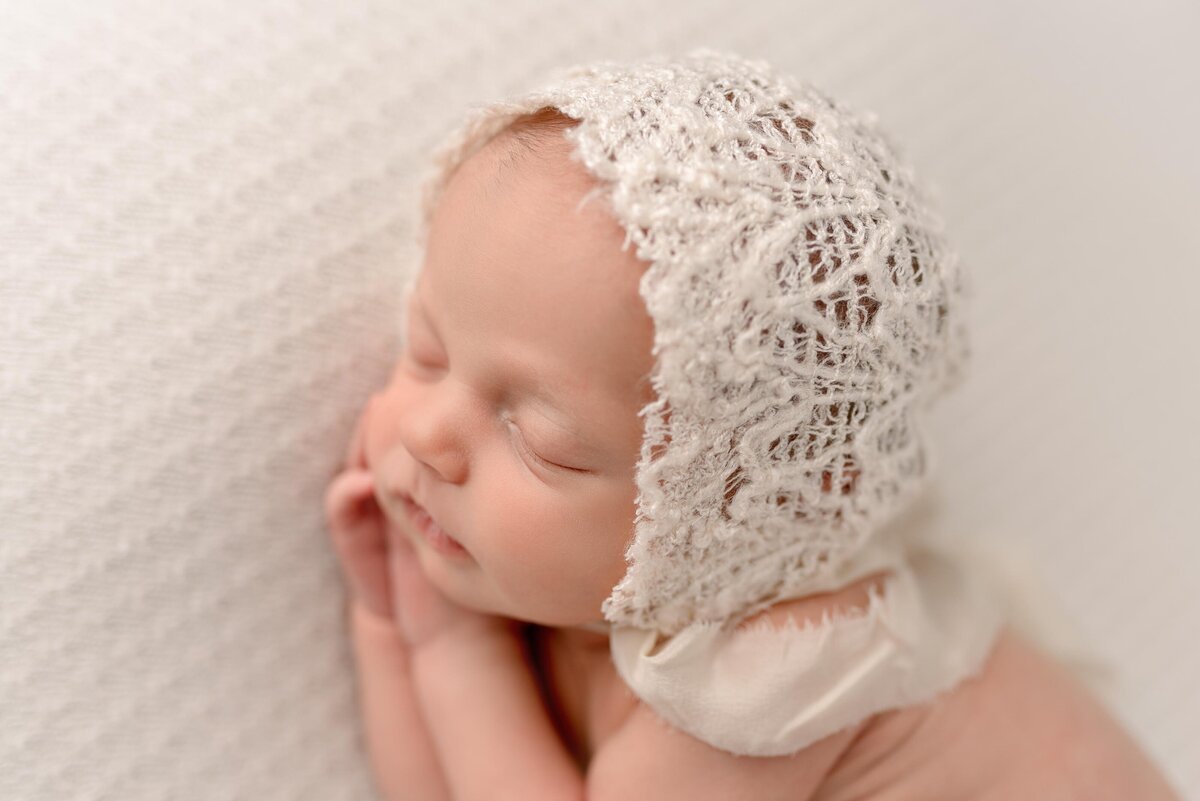Newborn baby girl laying on her side wearing a white lace bonnet
