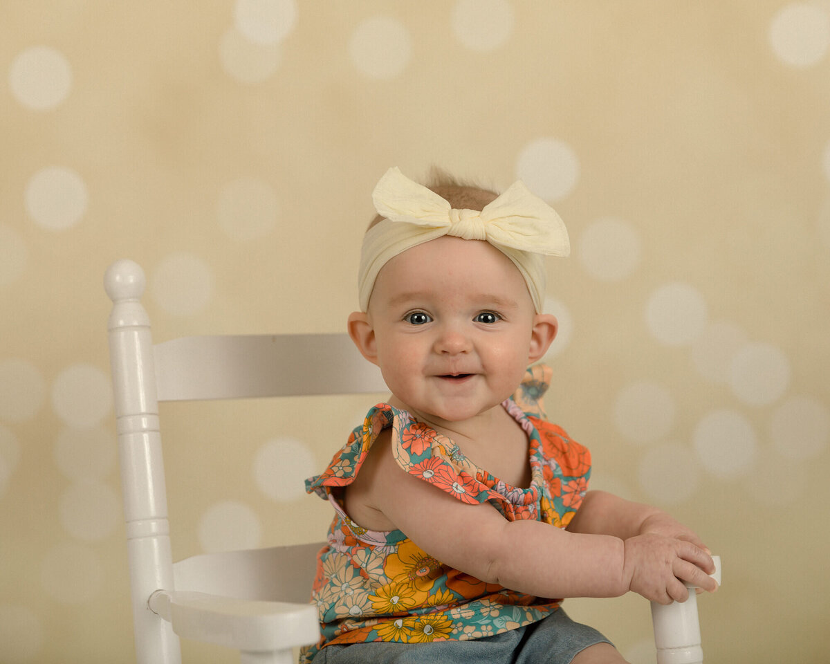 Baby girl with large white bow headband sitting in white rocking chair.