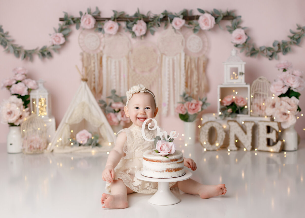 BoHo and floral cake smash in West Palm Beach and Wellington Florida. Baby girl is sitting with a cream lace romper with headband to match sitting next to a naked cake. In the background is a soft pink backdrop with hanging dream catchers and rose floral banner. There is a canvas lace tent with soft pink florals everywhere and a wood ONE.