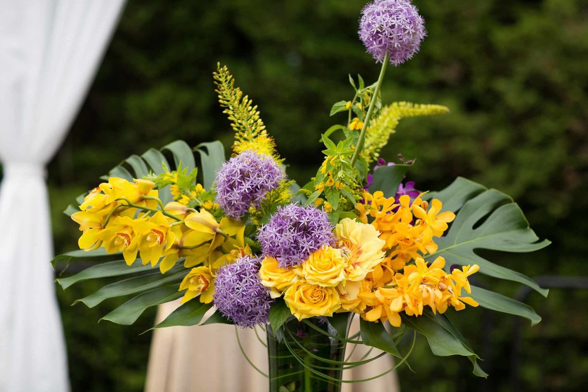 large flower arrangement of monstera leaves, large purple allium, yellow orchids, yellow eremurus, and yellow roses