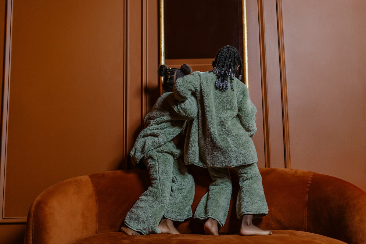 Modern and playful image of two young sisters naturally captured having fun in front of a mirror during a magazine-style photoshoot with a family photographer from the DFW area. The girls are dressed in matching cozy outfits, creating a contrasting and joyful atmosphere in this candid moment.