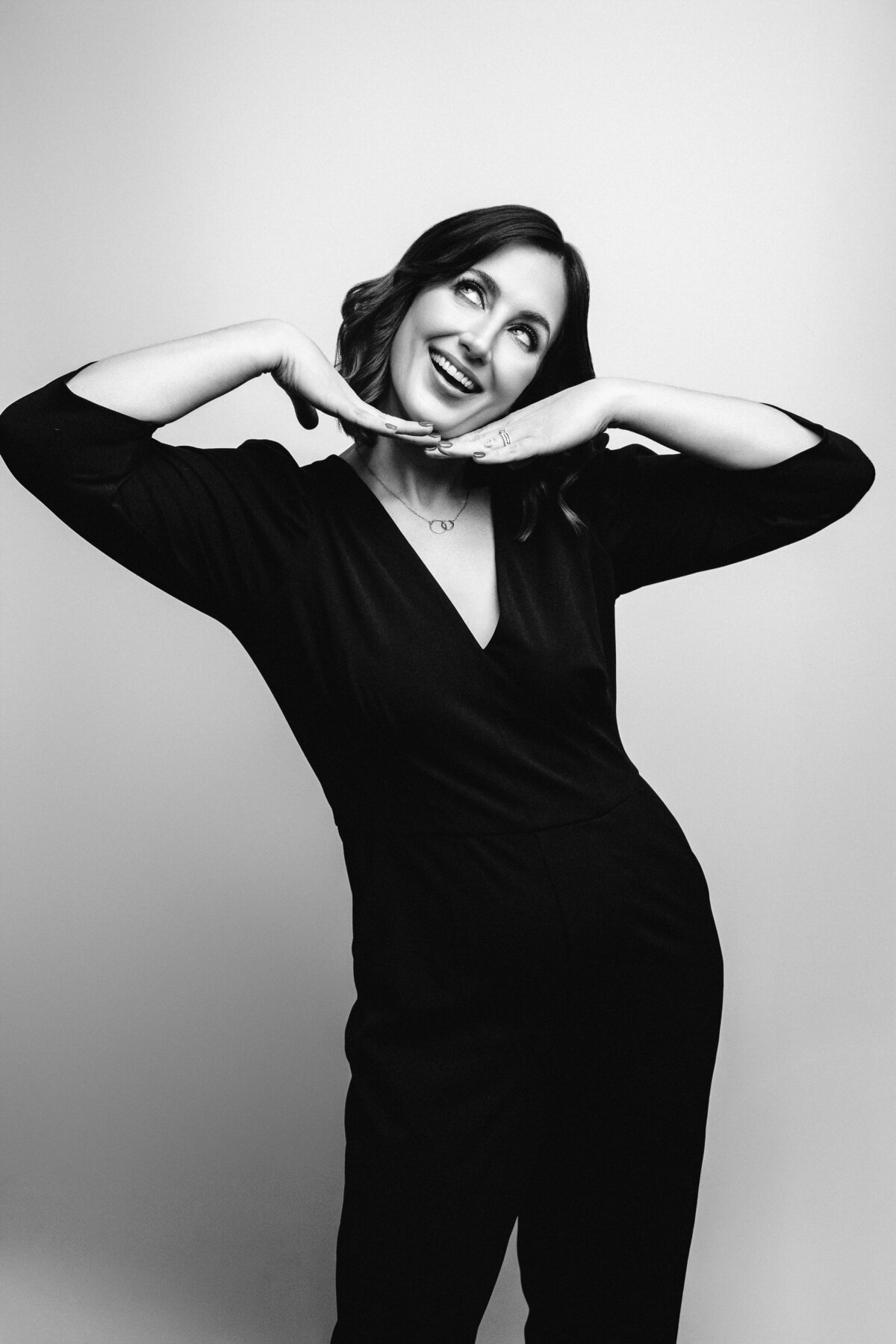 Black and white image of woman looking up and smiling with hands under her chin wearing a black jumpsuit on a white backdrop.