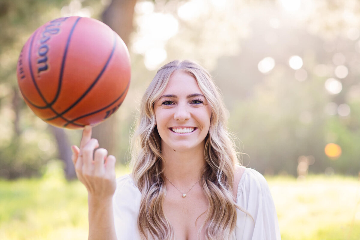 A woman with long blonde hair smiles while spinning a basketball on her finger. She is outdoors, surrounded by greenery and soft sunlight filtering through trees.