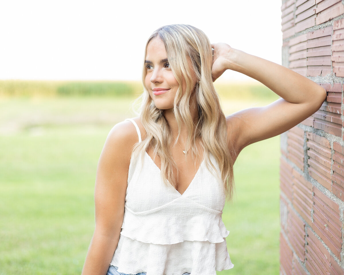 high school senior female with long blonde hair, standing in the grass, leaned against a brick wall, looking away from the camera with a soft smile, wearing a white spaghetti strap top