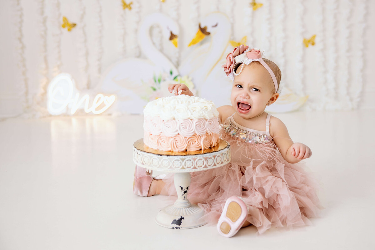 In Jacksonville, a baby in a pink dress and headband sits on the floor, joyfully smiling at a cake topped with pink and white swirls. The backdrop features swans and butterflies, with a neon "One" sign glowing. Capture these precious moments with Jacksonville cake smash photography.