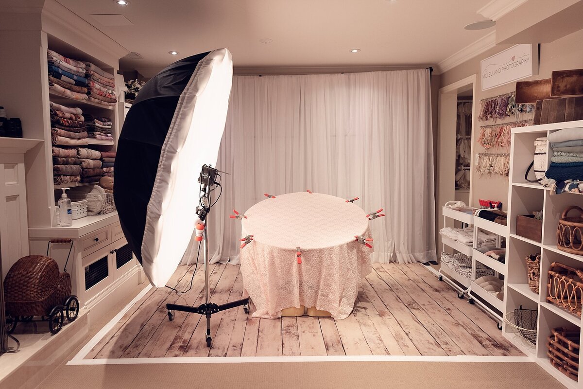 Professional newborn photography shooting space with studio lighting, posing table, wood floors, and organized props on open shelves.